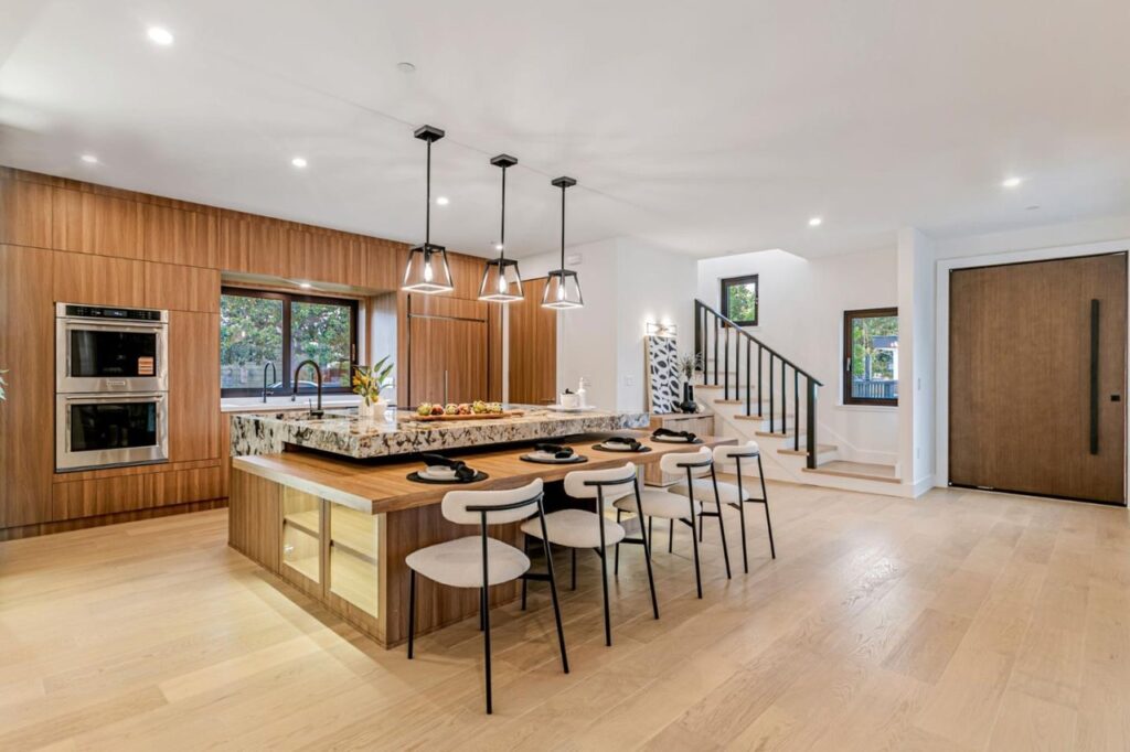 Modern kitchen and entryway interior at Hawthorne in Campbell, featuring a large marble and wood kitchen island with integrated seating, luxurious floor-to-ceiling wooden cabinetry, light wood flooring, and a staircase leading upstairs.
